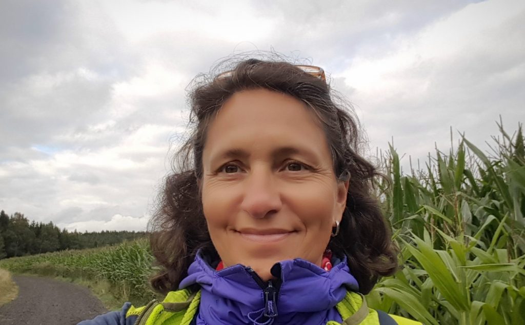 Ein Selfie von Gisela Jung auf einem Feldweg, rechts neben ihr ein Maisfeld.