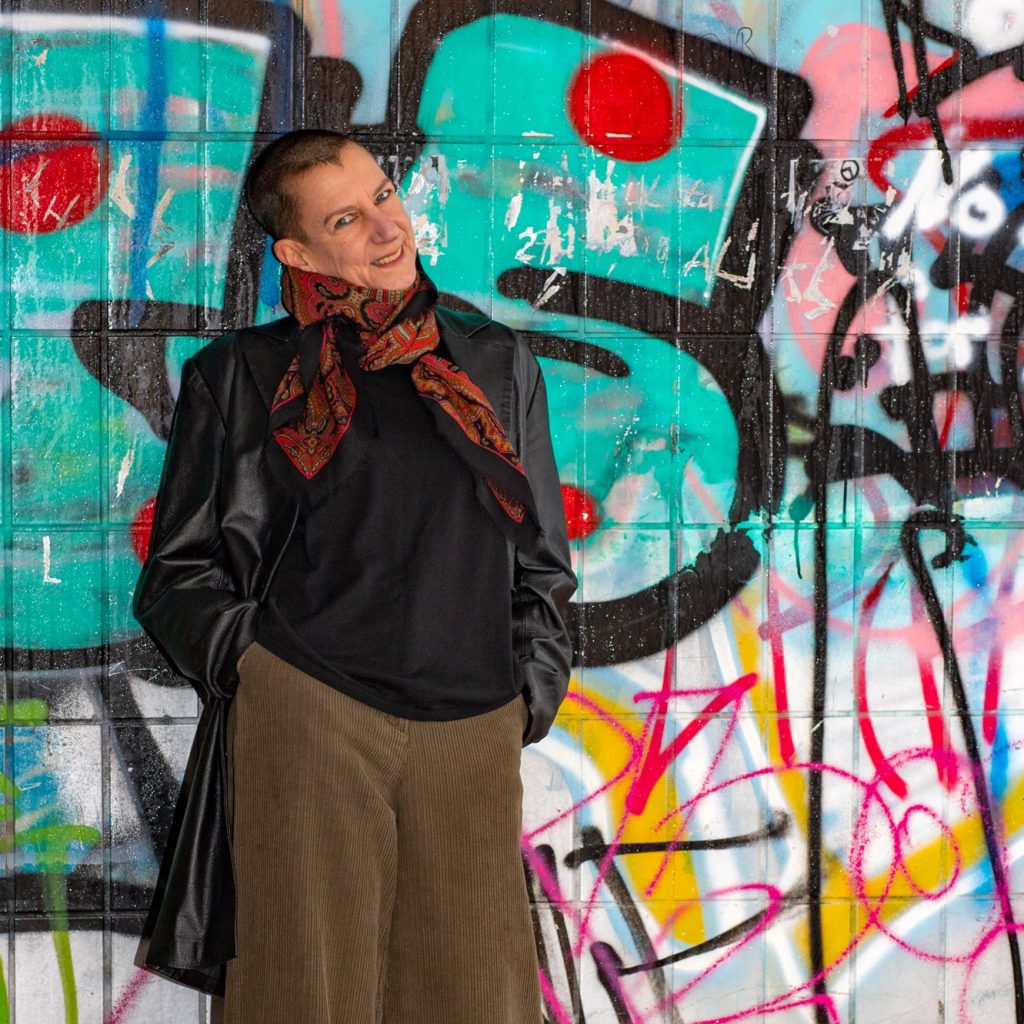 Rückkehr ins Leben: Sabine Scholze steht im Ledermantel vor einer Graffiti-Wand, die Hände in den Hosentaschen und lächelt.