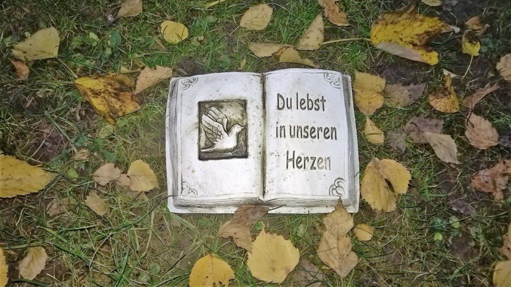 Eine Antwort auf die Frage "Was ist Trauer? " wird durch dieses Bild illustriert: Ein kleiner Grabstein in Form eines aufgeschlagenen Buches, zwischen welken Blättern, auf dem steht: "Du lebst in unseren Herzen."