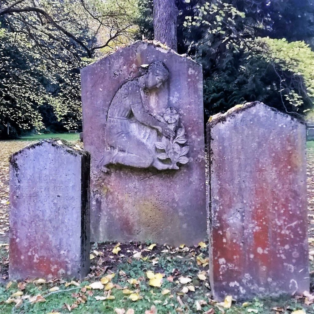 Ein weiteres Foto vom Göttinger Stadtfriedhof: Drei Grabsteine als Triptychon, in der Mitte eine Frauengestalt, die eine Blume berührt.