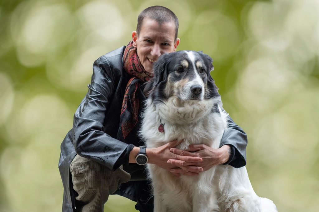 Selbstliebe bei der Fotosession: Sabine Scholze in der Hocke neben ihrem schwarz-weißen Herdenschutzhund vor einem grünen Hintergrund.